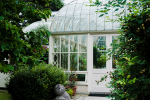 A bright glass conservatory surrounded by lush green plants and shrubs in a garden, with sunlight reflecting off the curved roof and white-framed windows.