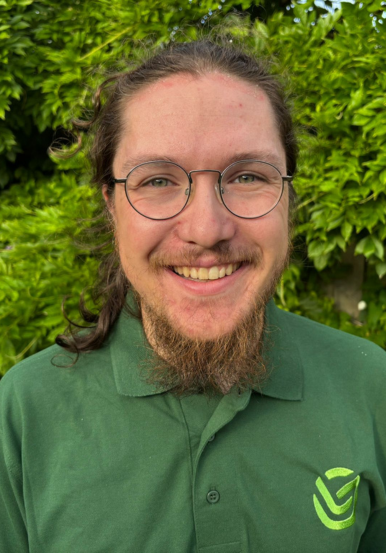 Headshot of Jude DeSouza, Environmental Consultant at Little Green Consulting, smiling and wearing a green Little Green Consulting polo shirt, pictured outdoors against a leafy green background.